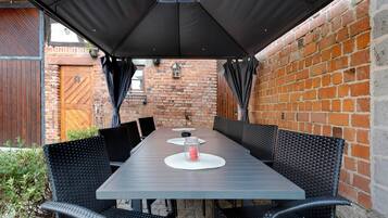 Une terrasse extérieure avec une longue table, des chaises en osier noir et un parasol noir offrant de l’ombre.