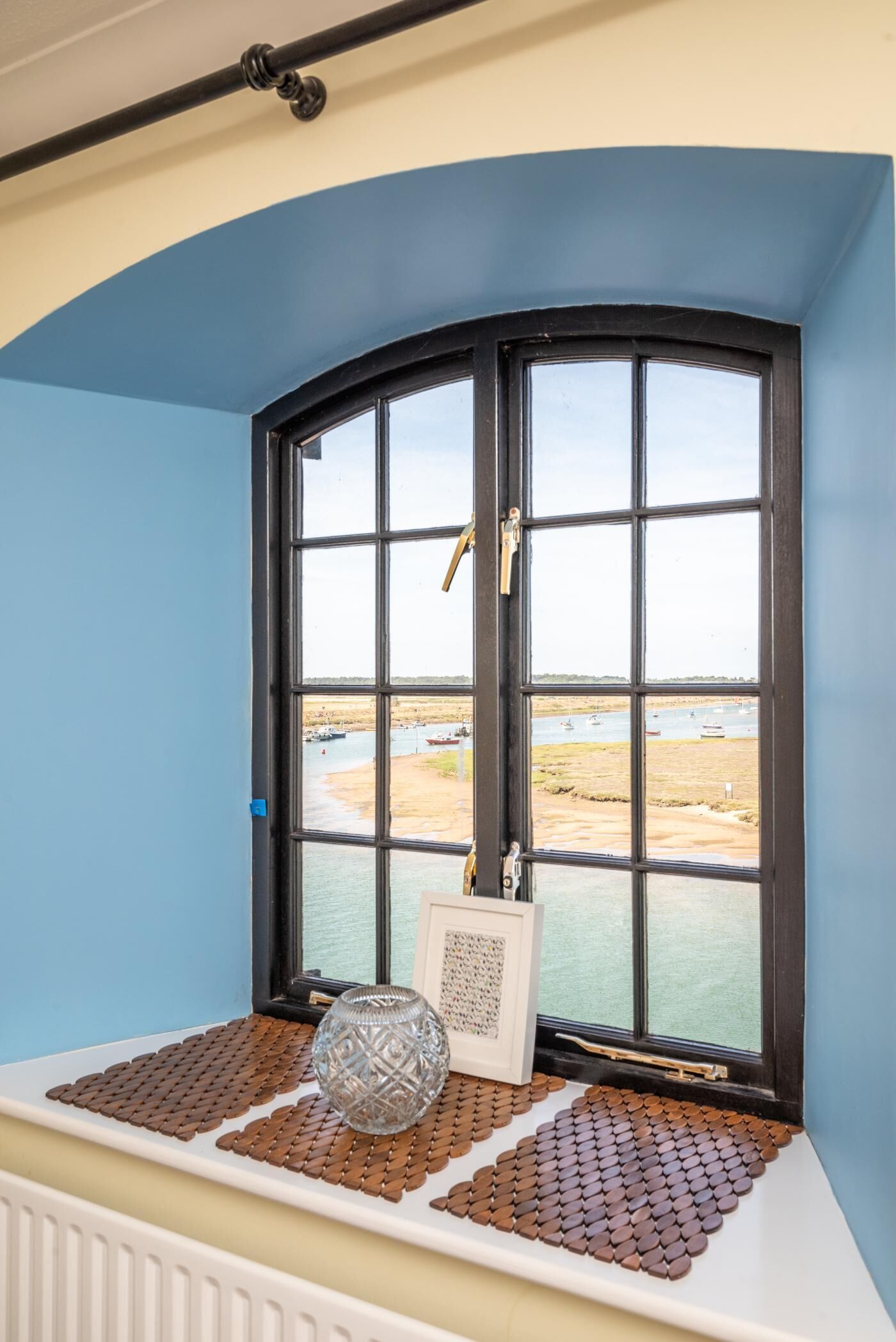 A bay window with a view of a water body and boats, a decorative glass vase, and a framed picture on a textured mat.
