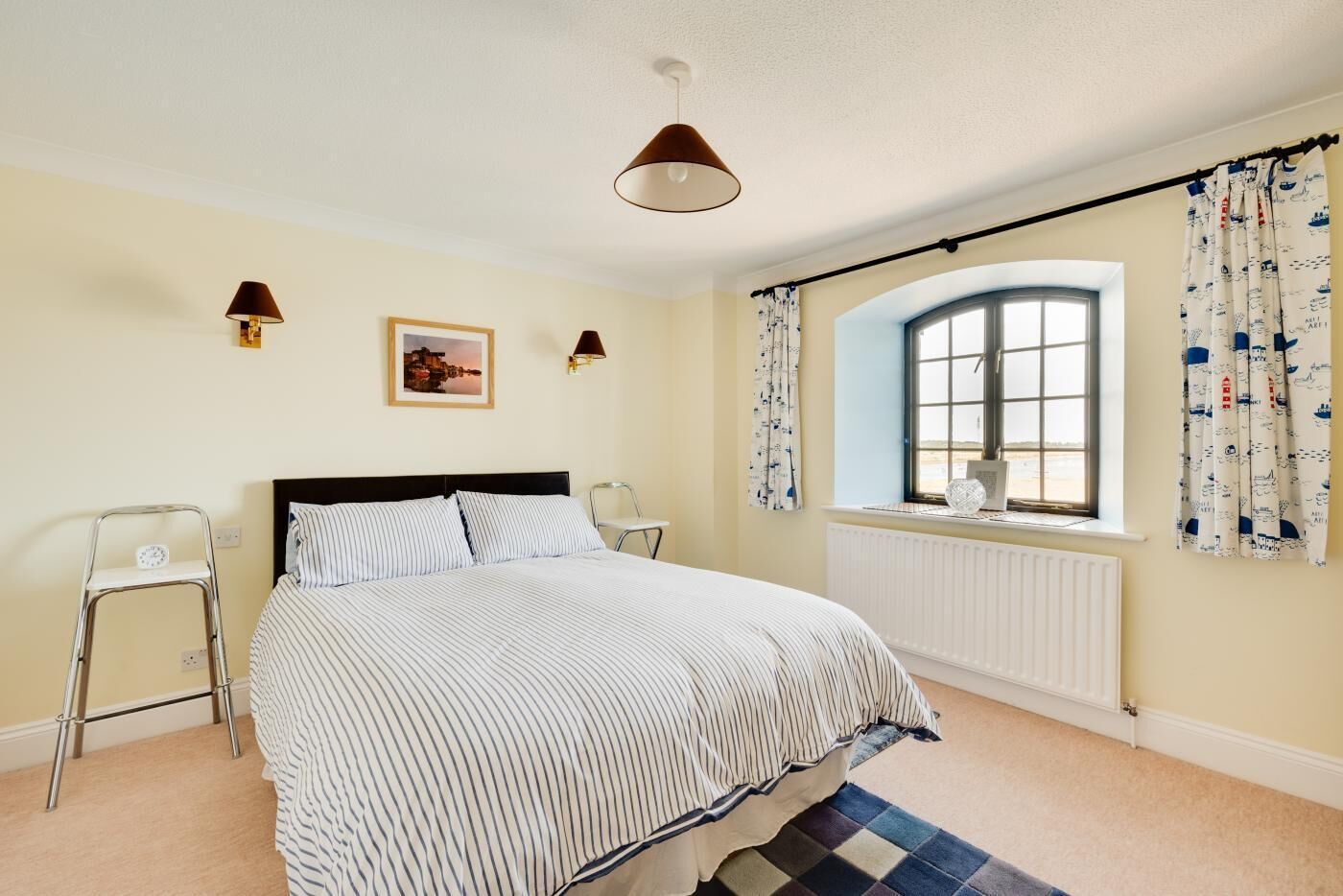 A bedroom with a bed, two bedside lamps, a window with curtains, a radiator, and a small picture on the wall.