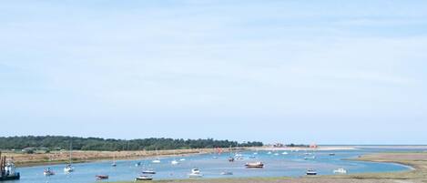 A coastal landscape with boats anchored in a calm bay, a sandy beach, and a distant shoreline with vegetation.