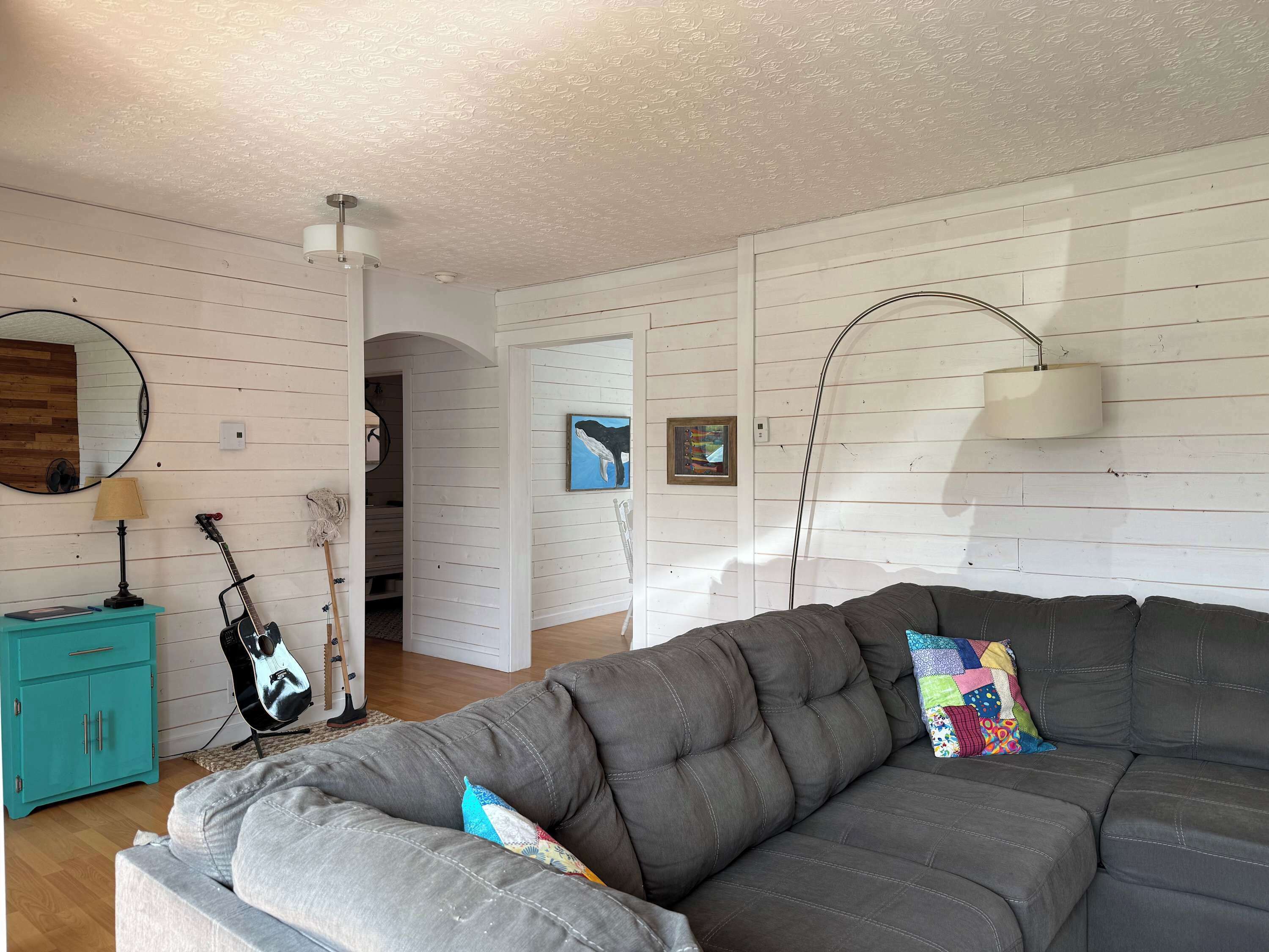 A living room with a grey sofa, a teal cabinet, a guitar, a lamp, and a mirror on the wall.