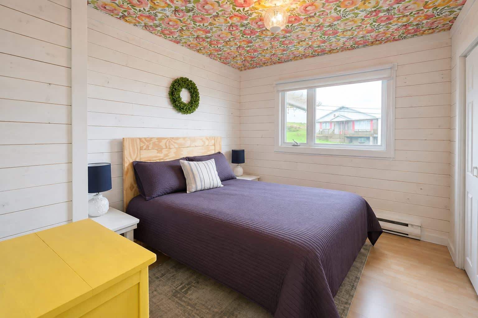 A bedroom with a wooden headboard, a purple bedspread, a yellow nightstand, a white lamp, and a window with a view of houses.