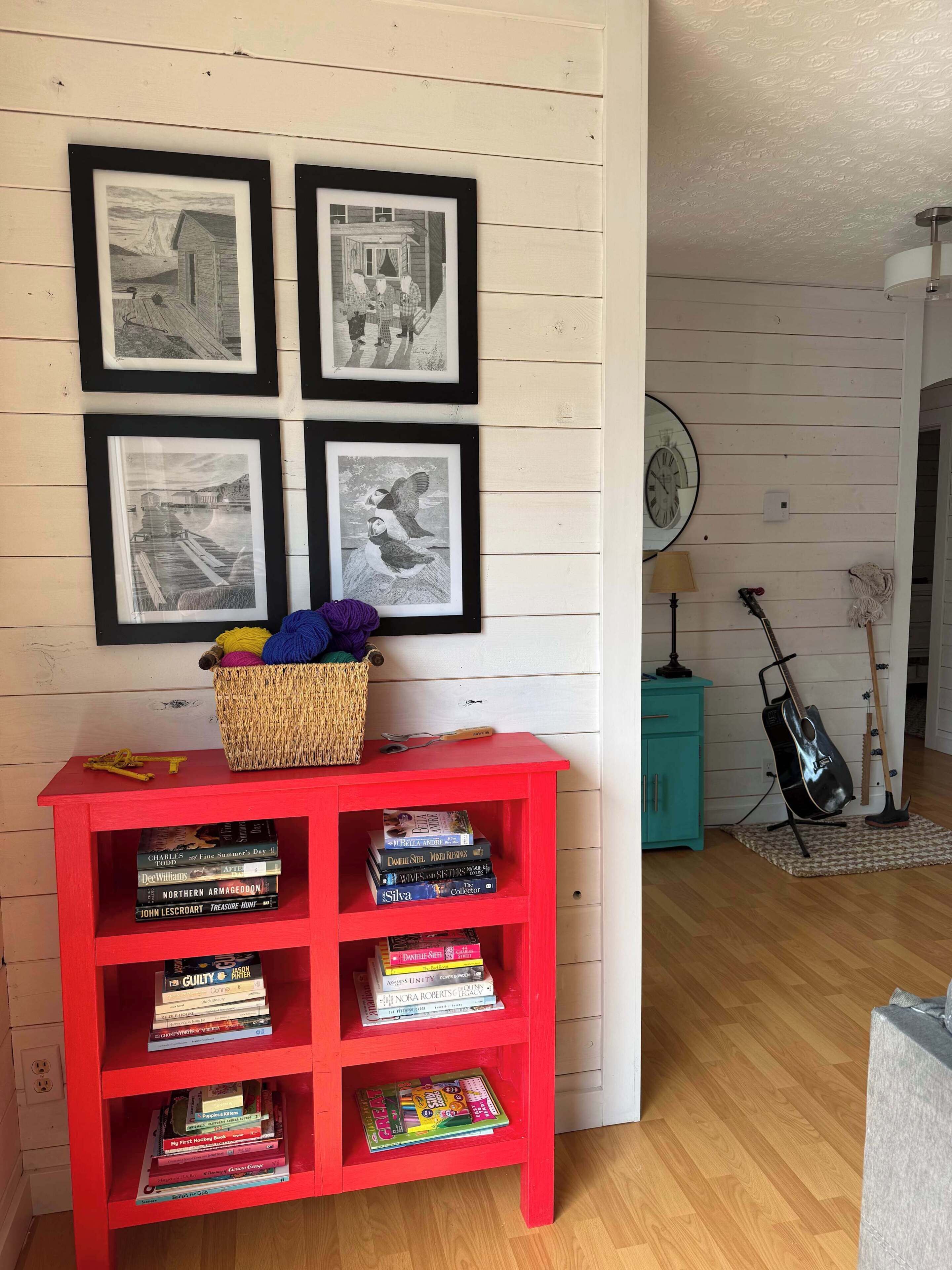 Een kamer met een rode boekenkast vol boeken, een rieten mand met wol en vier zwart-wit foto's in lijsten aan de muur.
