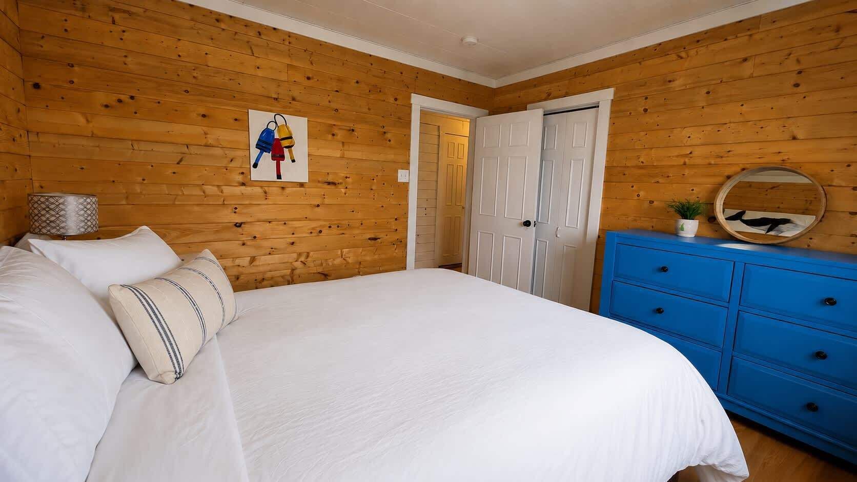 Een slaapkamer met een houten lambrisering, een bed met witte lakens, een blauwe dressoir en een deur.