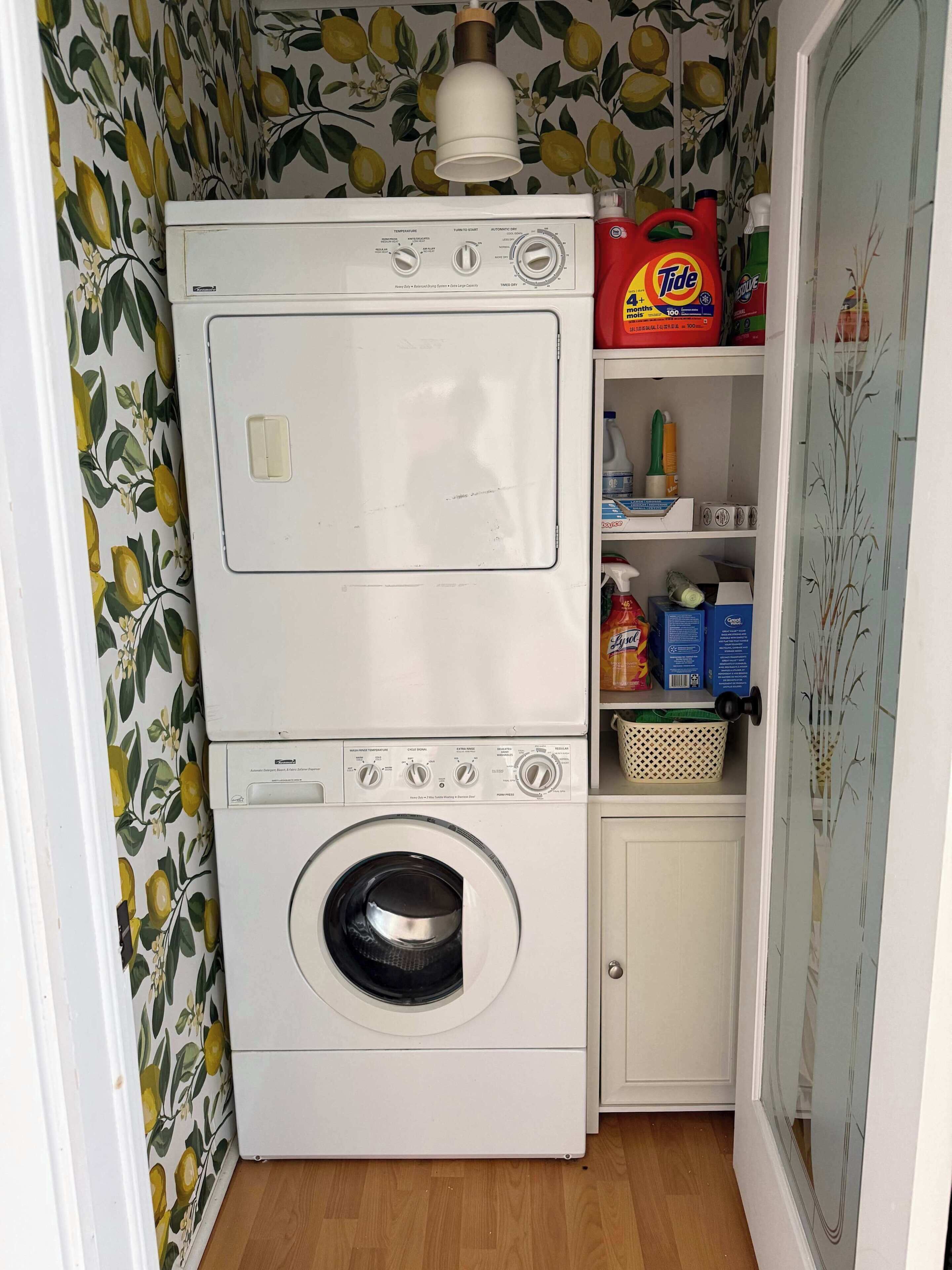 A compact laundry area with a washing machine and dryer, a shelf with cleaning supplies, and a patterned wall.