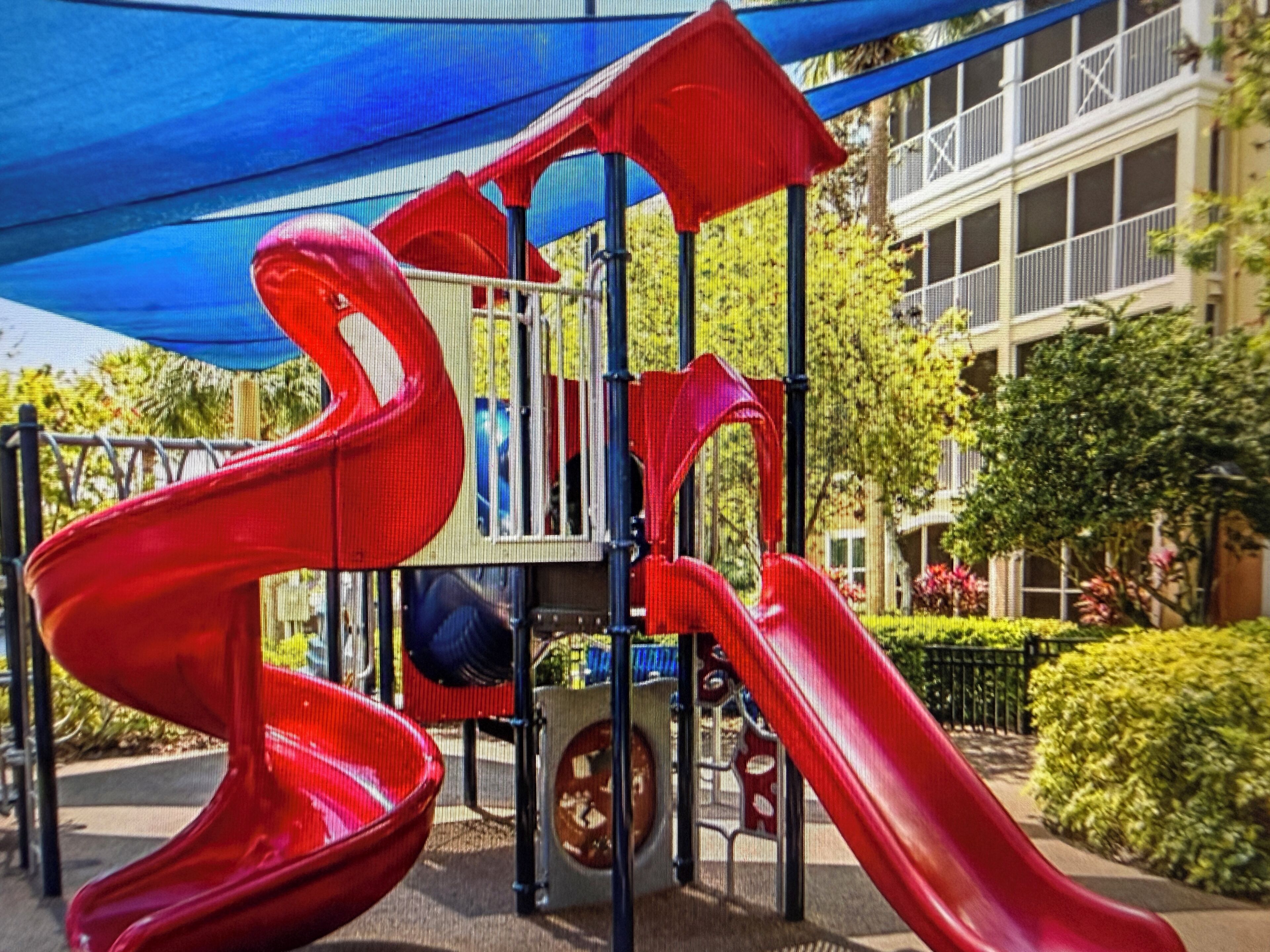 Une aire de jeux avec des toboggans rouges et une structure bleue en forme de tonnelle.