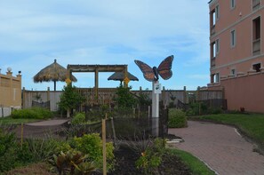 Un jardín con una escultura de mariposa, una estructura con techo de paja y un camino pavimentado.