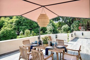 Uma área de refeições ao ar livre com uma mesa posta para quatro pessoas, rodeada de cadeiras, sob um grande guarda-sol.