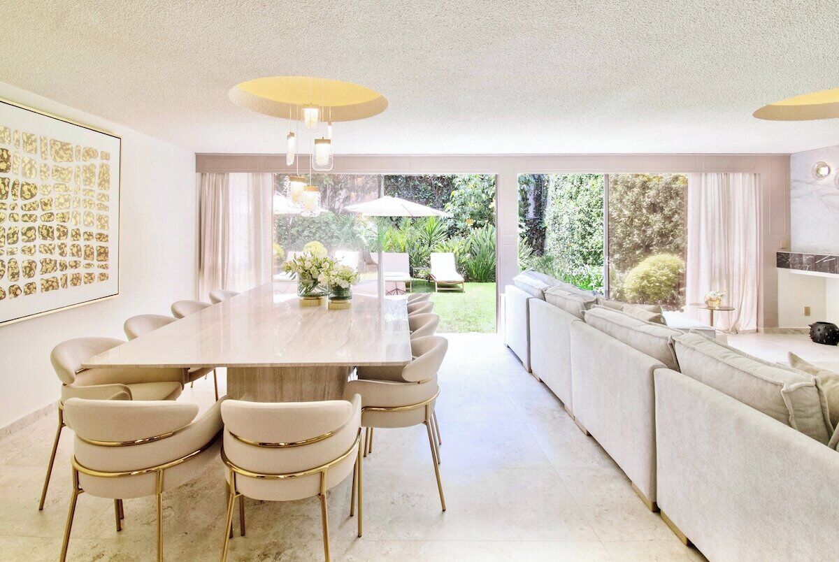 A modern dining area with a long table and chairs, a sofa, and a view of the garden.