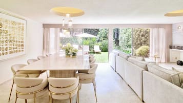 A modern dining area with a long table and chairs, a sofa, and a view of the garden.