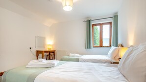 A hotel room with two beds, a table, a lamp, and a window with curtains. - 3 Bed in Kilkhampton (oc-l27383) (Kilkhampton)