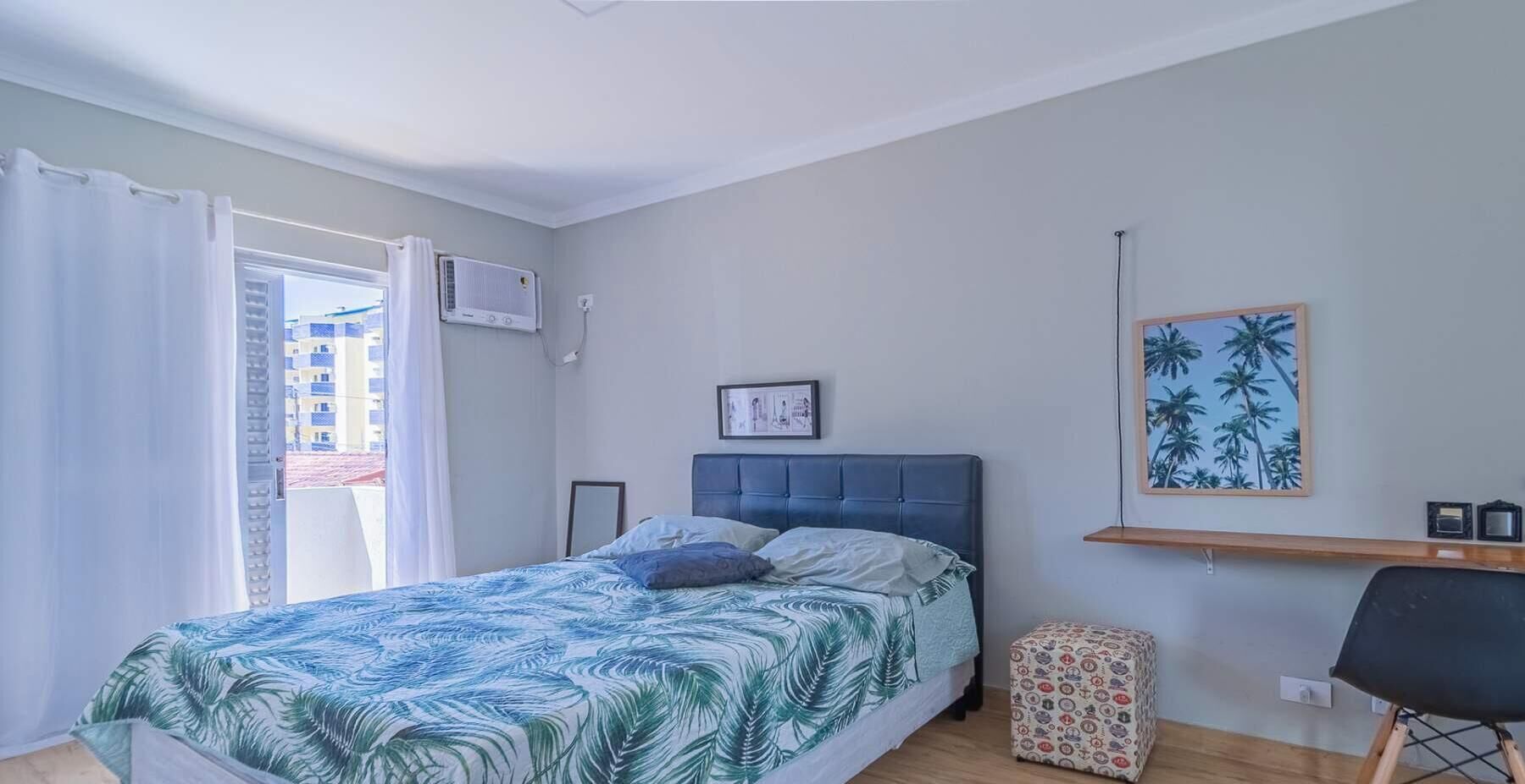 Ein Schlafzimmer mit einem Bett, einem Fenster mit Vorhängen, einer Wandklimaanlage, einem gerahmten Bild, einer kleinen Ablage und einem gemusterten Hocker.