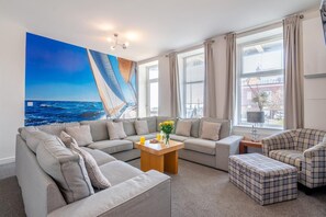 Ein modernes Wohnzimmer mit einem großen Gemälde einer Segelyacht an der Wand, einem grauen Sofa, einem karierten Sessel und einem kleinen Holztisch mit Blumen in einer Vase.