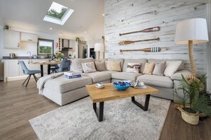 Un salon moderne avec un canapé d’angle, une table basse et un coin repas, la cuisine étant visible au fond.