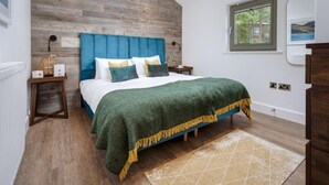 A bedroom with a wooden headboard, a bed with a green blanket, a bedside table with a lamp, and a window with a view of trees. - 2 Bed in Newby Bridge (oc-b30231) (Newby Bridge)