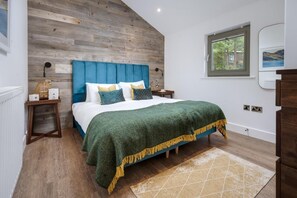 Ein Schlafzimmer mit einem hölzernen Kopfteil, einem Bett mit grüner Decke, einem Nachttisch mit Lampe und einem Fenster mit Blick auf Bäume.