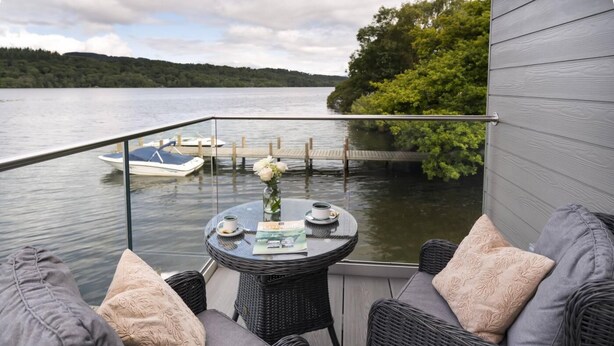 A balcony with wicker furniture, a glass table, and a view of a lake with a dock and boat. - 2 Bed in Newby Bridge (oc-b30231) (Newby Bridge)