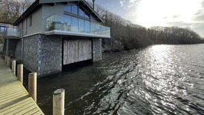 A modern houseboat with a glass balcony, wooden deck, and stone facade, situated on a calm body of water. - 2 Bed in Newby Bridge (oc-b30231) (Newby Bridge)