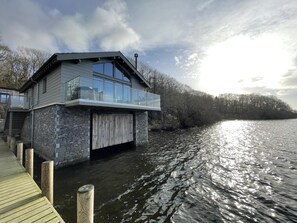 Ein modernes Hausboot mit Glasbalkon, Holzterrasse und Steinaußenwand, gelegen auf ruhigem Gewässer.