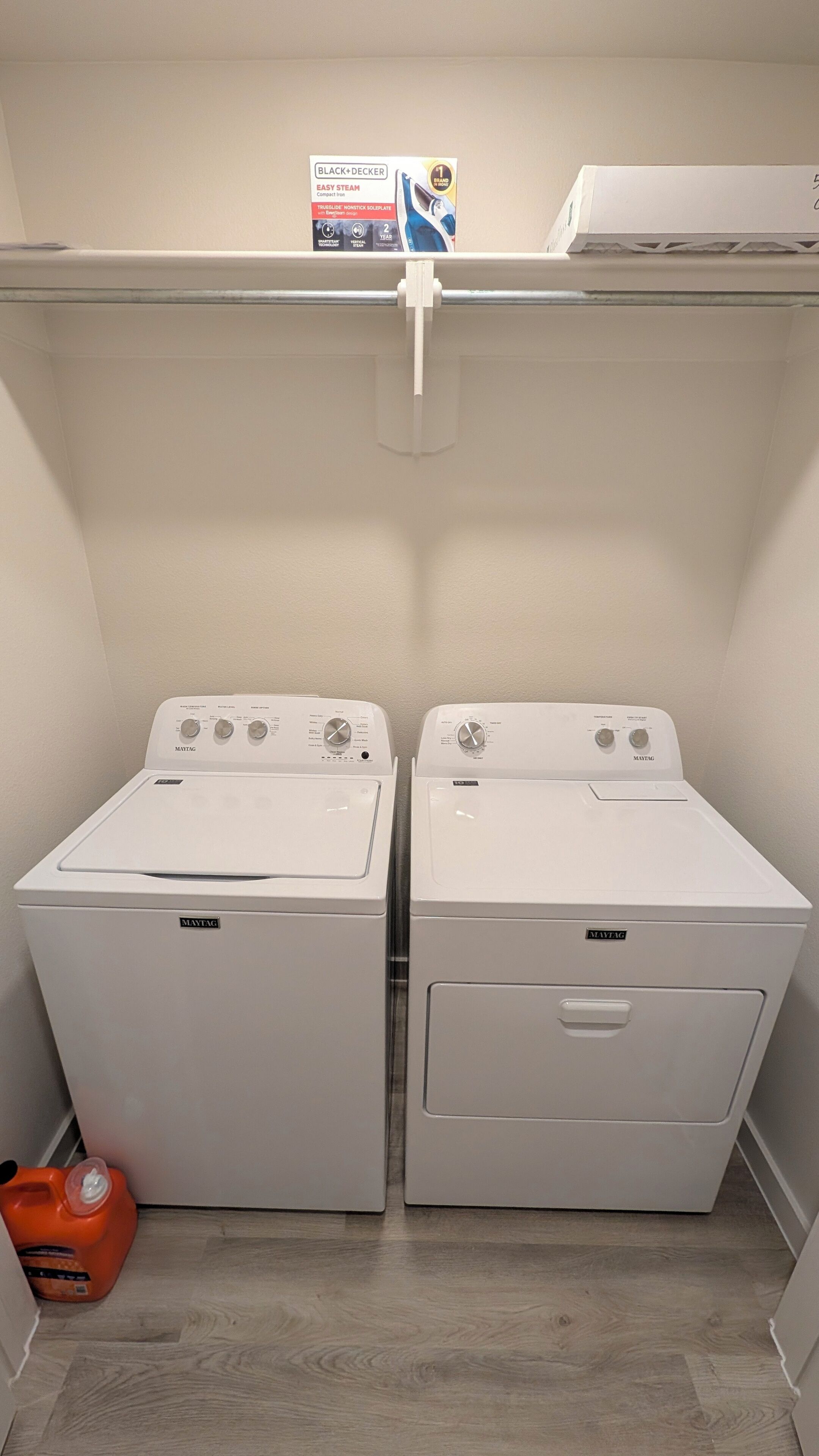 Una lavandería con dos lavadoras y secadoras Maytag, una botella de detergente y un juego de plancha Black+Decker sobre la estantería.