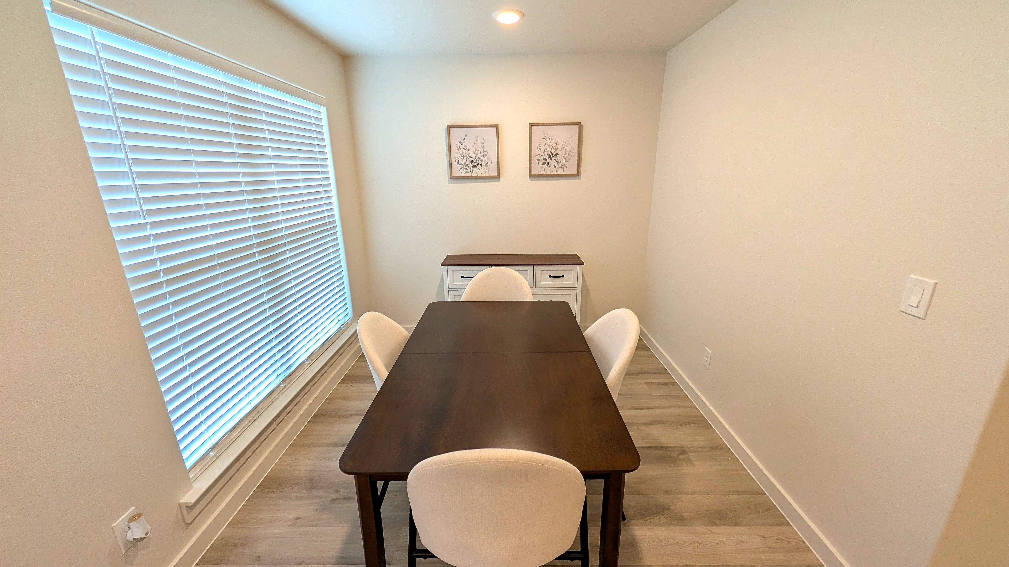 Un espace repas comprenant une longue table, des chaises, une fenêtre avec des stores, un luminaire fixé au mur et deux tableaux encadrés.