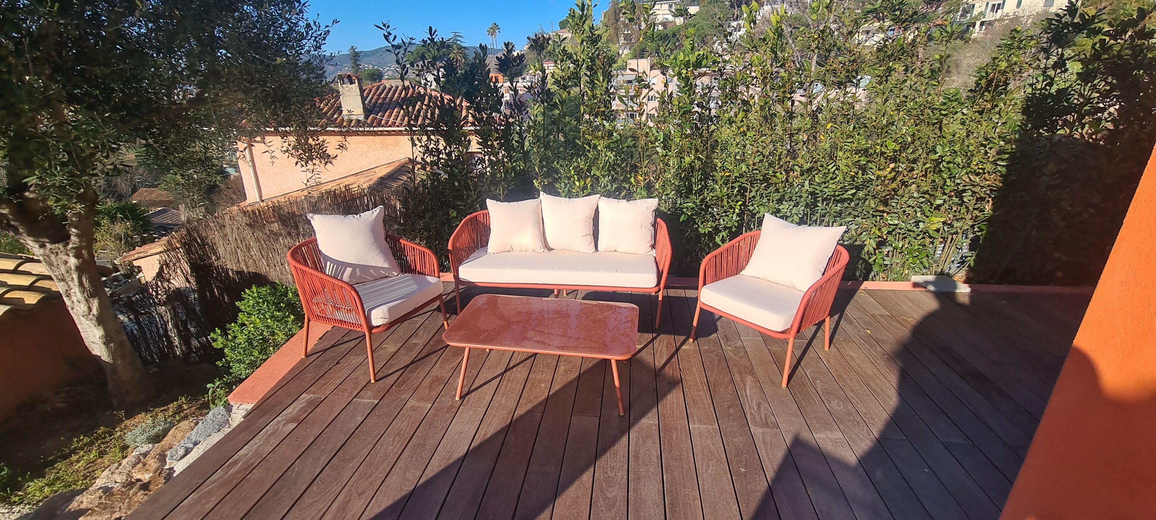 A wooden deck with outdoor furniture, including a sofa and chairs, surrounded by greenery and buildings in the background.