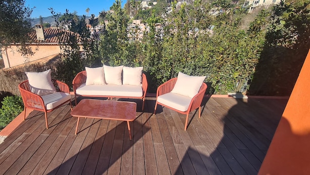 Una terraza de madera con mobiliario exterior, incluyendo un sofá y sillas, rodeada de vegetación y edificios al fondo.