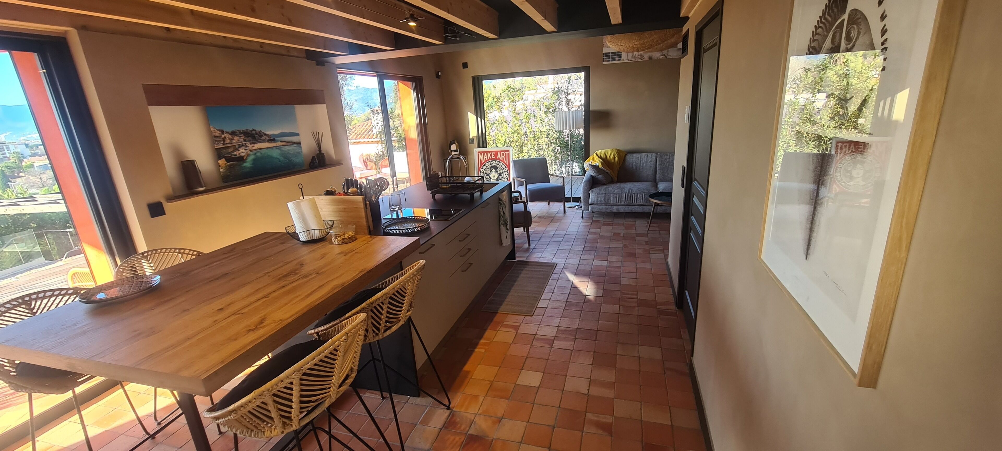 A modern kitchen with a wooden countertop, wicker chairs, and a view of a coastal landscape through large windows.