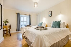 A hotel room with two beds, a desk with a chair, a window with curtains, and a painting on the wall. - 2 Bed in St. Issey (oc-c31993) (Tredinnick)