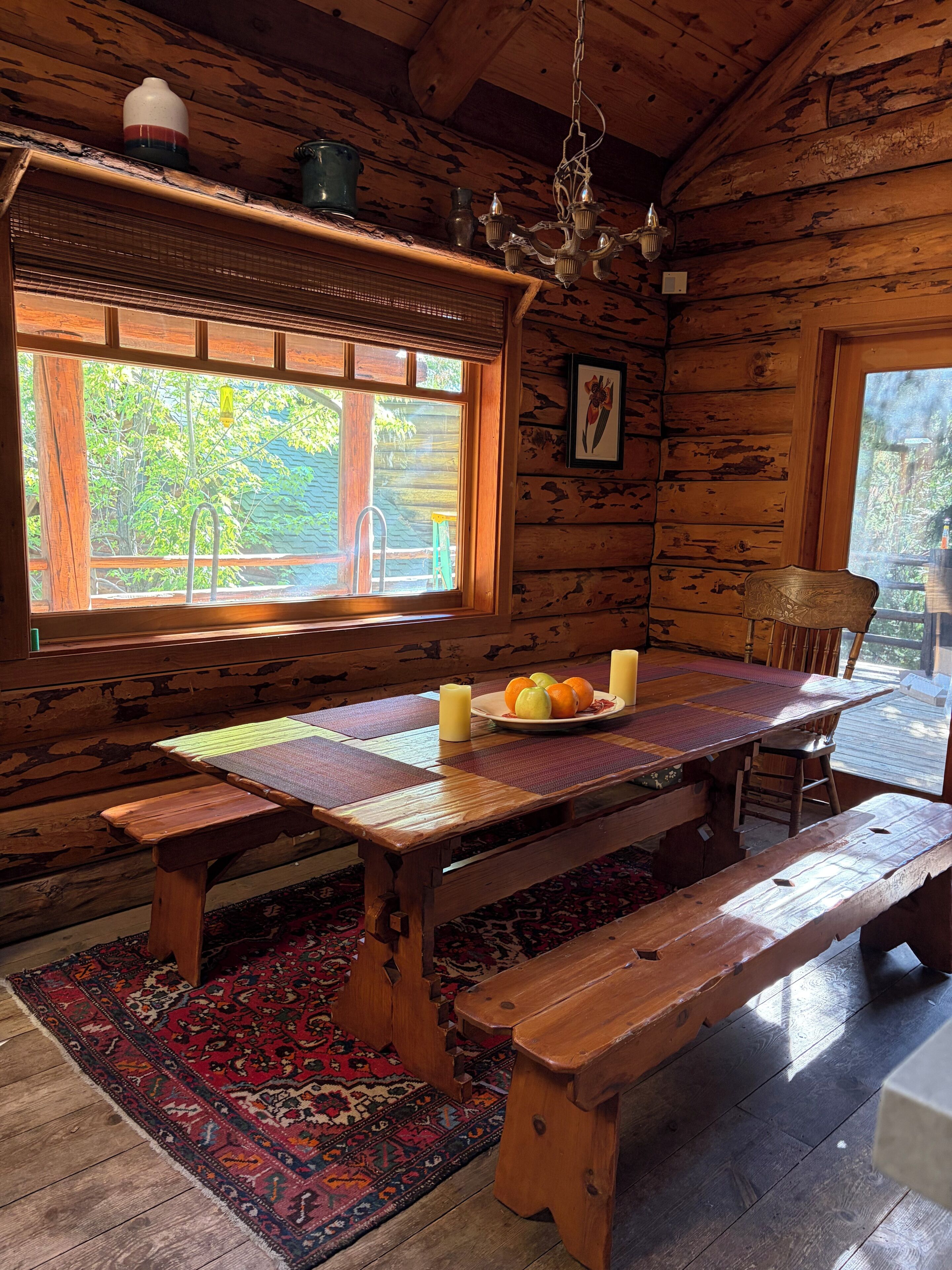 Interior de una cabaña de madera con una gran mesa de comedor, bancos, una araña y una ventana con vistas a árboles.