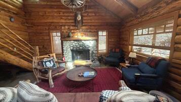 A log cabin interior with a stone fireplace, wooden furniture, and a ceiling fan.
