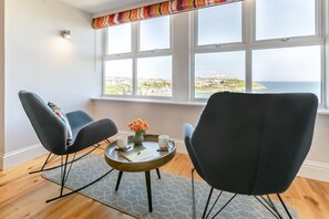 A room with two chairs, a small table with a vase of flowers, and a view of the sea through large windows. - 3 Bed in Newquay (oc-o32258) (Newquay)