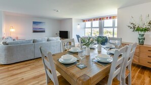 Uma sala de jantar com uma mesa de madeira posta para quatro, cadeiras brancas e um sofá ao fundo.