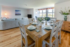 A dining area with a wooden table set for four, white chairs, and a sofa in the background. - 3 Bed in Newquay (oc-o32258) (Newquay)