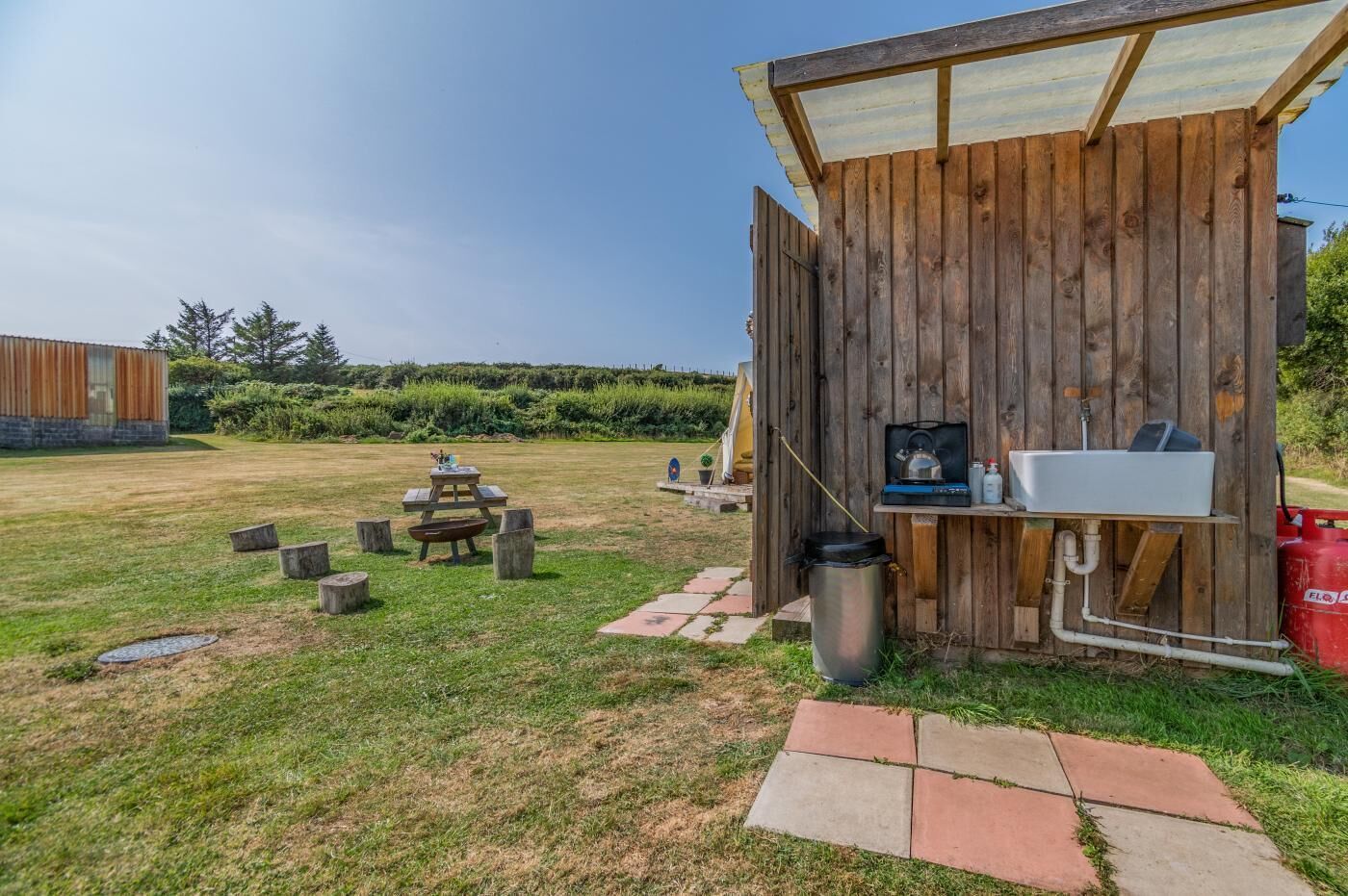 Een houten buitenkeuken met een barbecue en spoelbak, een picknicktafel en een vuilnisbak.