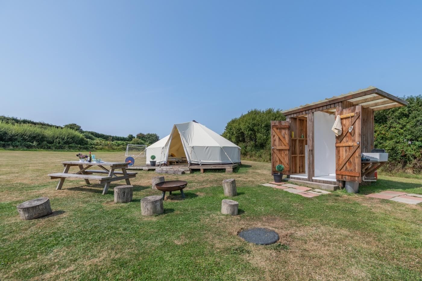 Un terrain de camping avec une tente, des toilettes sèches en bois, des tables de pique-nique et un champ d’herbe.