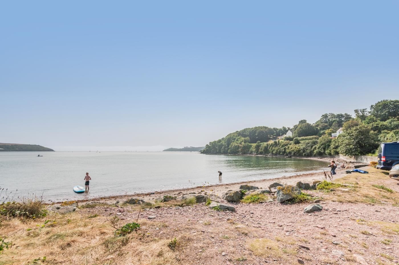 Een strand met een heldere lucht, een persoon die staat met een surfplank, en een voertuig geparkeerd aan de kust.