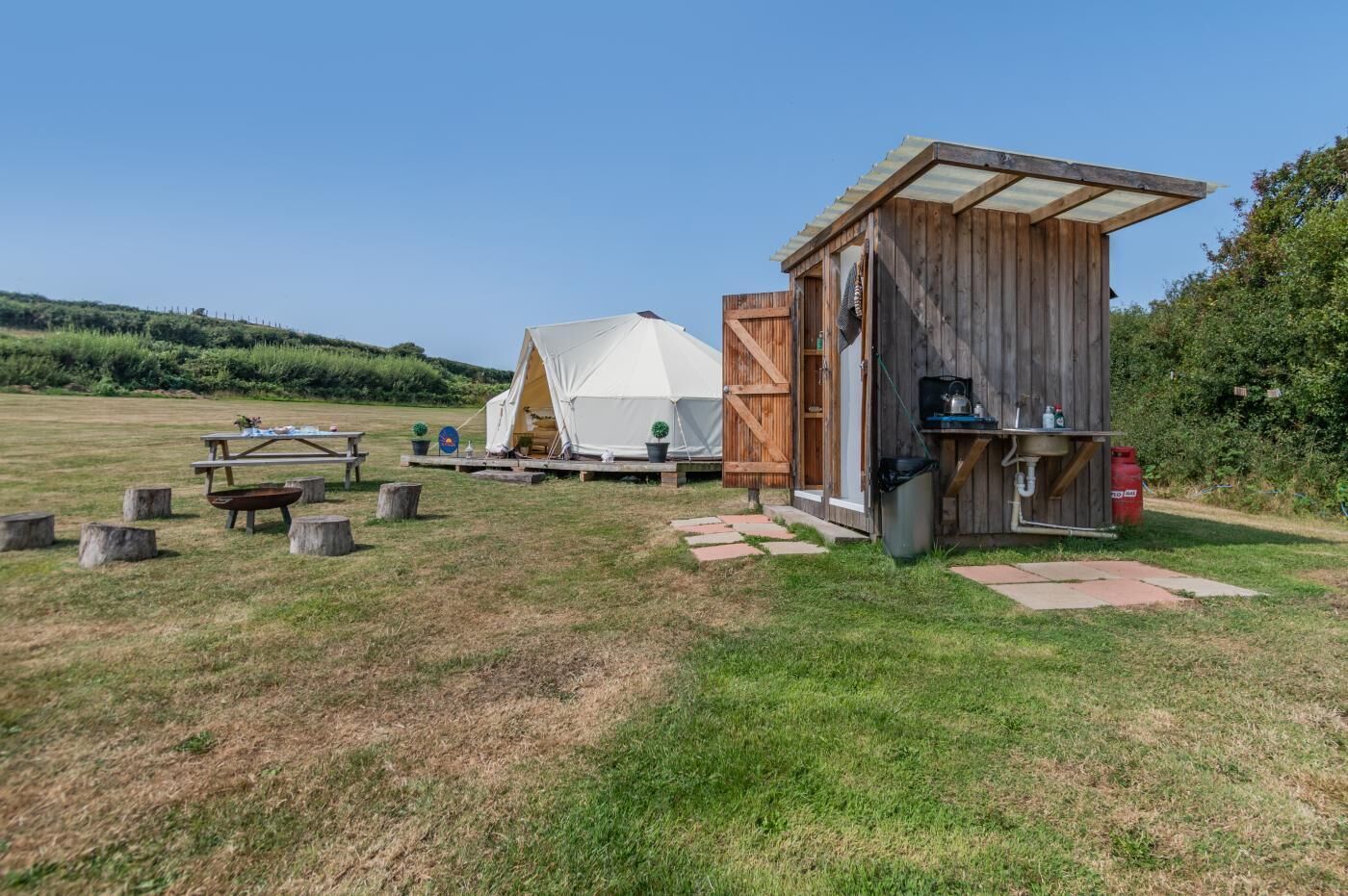 A campsite with a wooden structure, a tent, picnic tables, and a portable toilet.