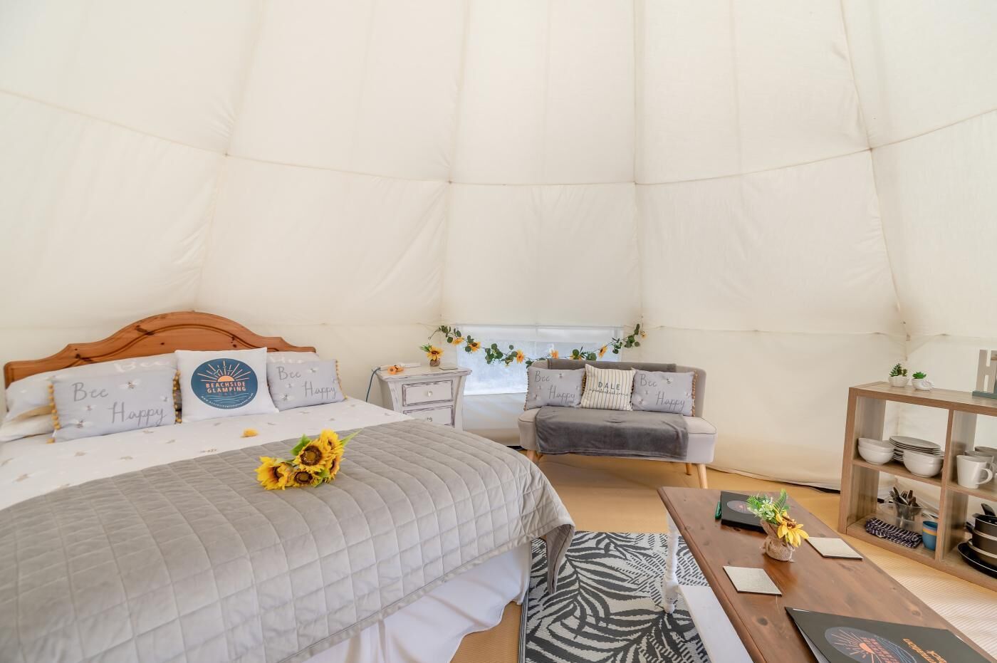 A tented accommodation room with a bed, a sofa, a wooden side table, and a small table with a vase.