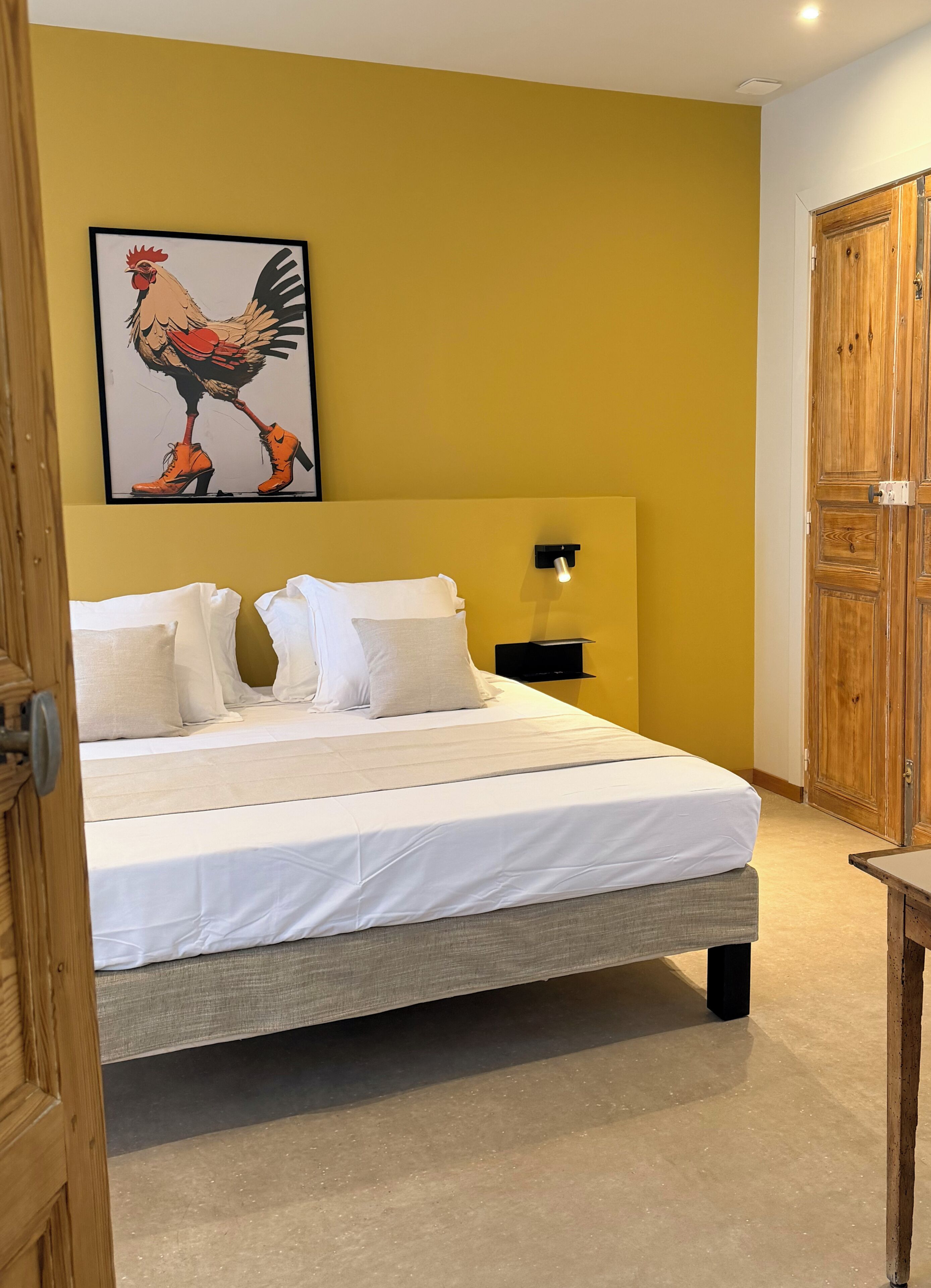 Een slaapkamer met een bed, kussens, een nachtkastje, een wandlamp en een houten deur.