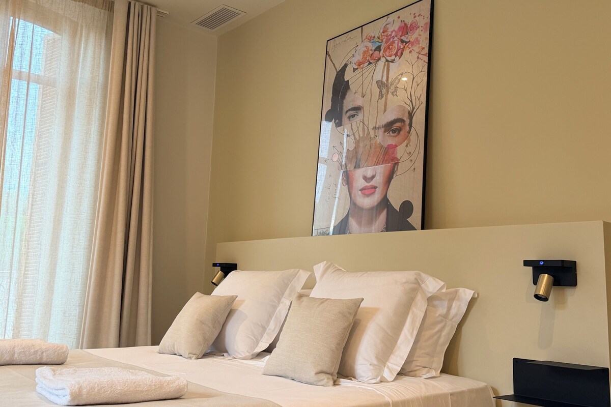 A hotel room with a bed, pillows, a headboard, a wall-mounted artwork, a window with curtains, and a wall-mounted light fixture.