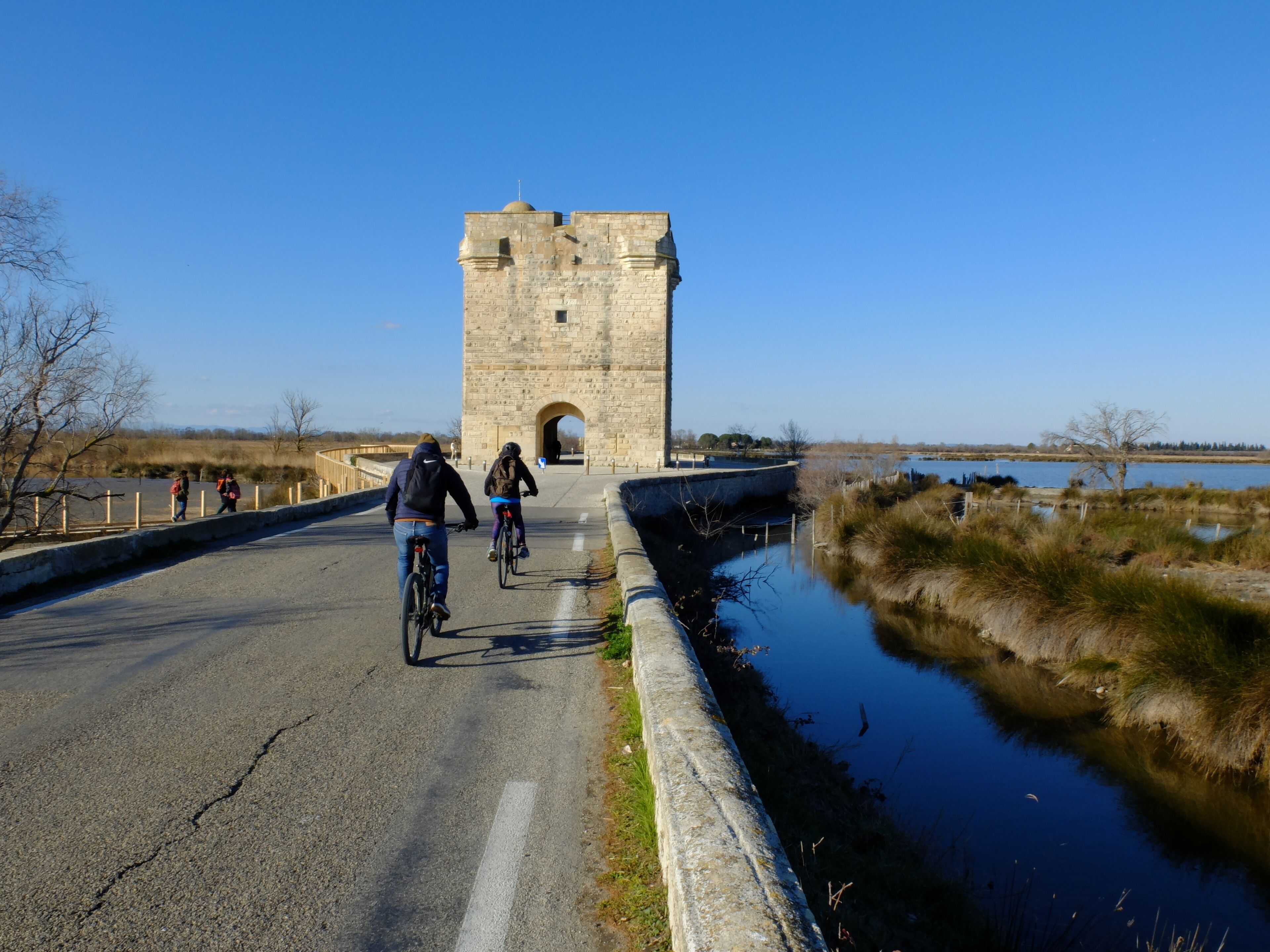 A stone tower with an arched entrance, a paved road beside a body of water, and cyclists on the road.