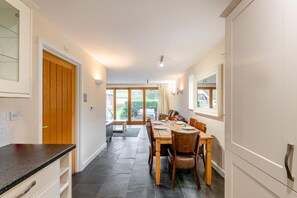 A modern kitchen with a dining area, featuring a long countertop, wooden cabinets, and a dining table set for four. - 3 Bed in Blackawton (oc-c32809) (Blackawton)