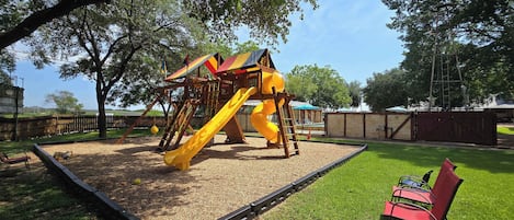 A playground with a slide and climbing structures, surrounded by a fenced area and trees.