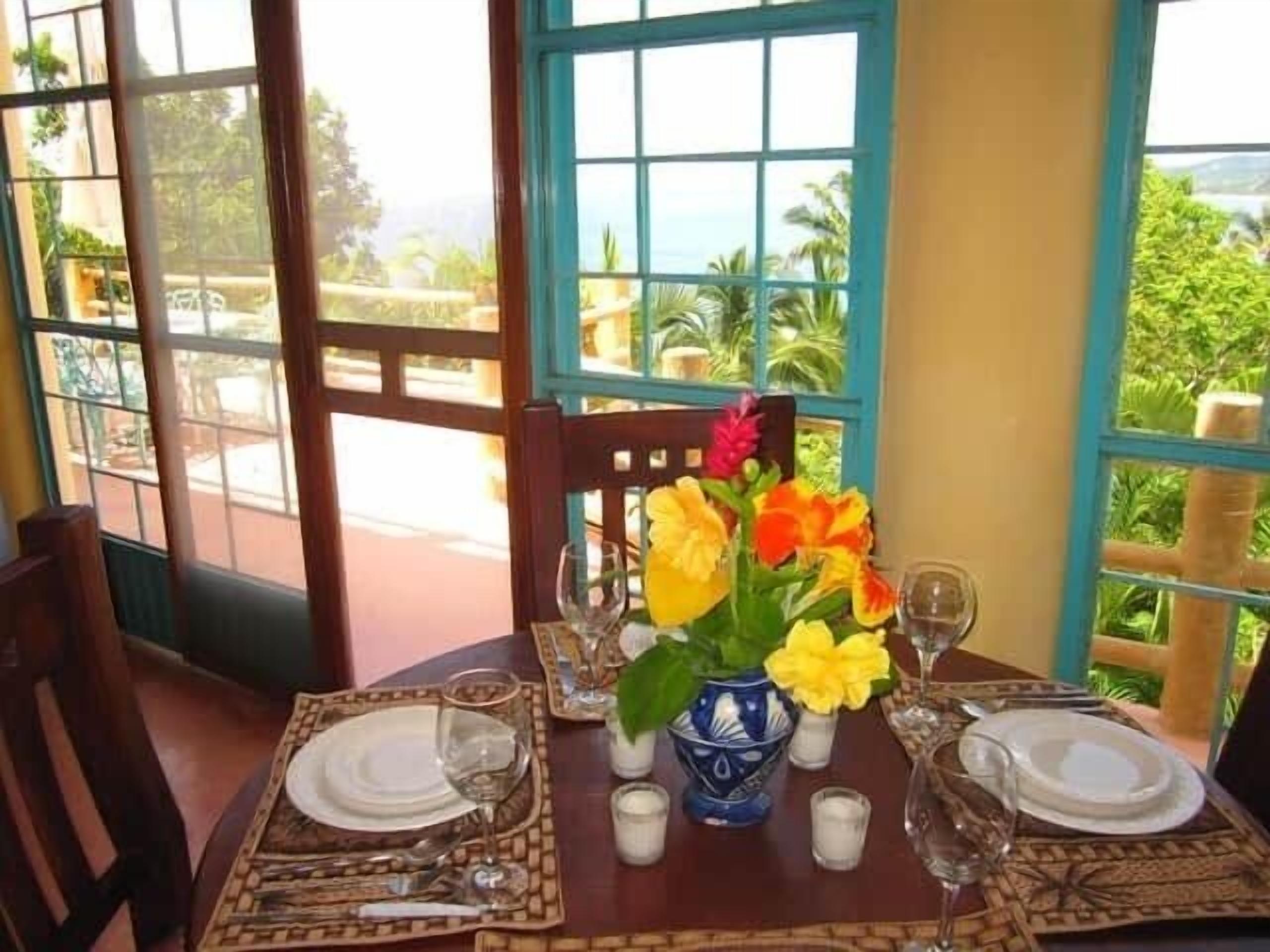 Mesa lista para comer, con un jarrón de flores, vista a follaje y un edificio al fondo.
