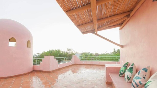 Eine Terrasse mit rosa Dach, Fliesenboden, einer Holz-Pergola und einer Reihe Kissen.