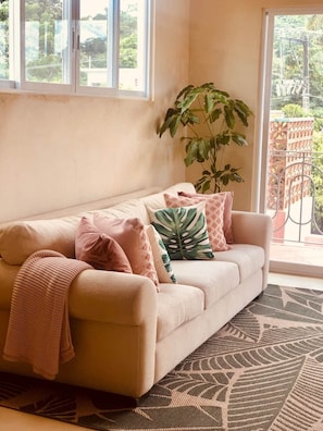 A beige sofa with a patterned throw blanket and multiple pillows, a large green plant, and a patterned rug in a room with large windows and a balcony. - Bella Rosa, center of town, steps to the beach. (San Francisco)