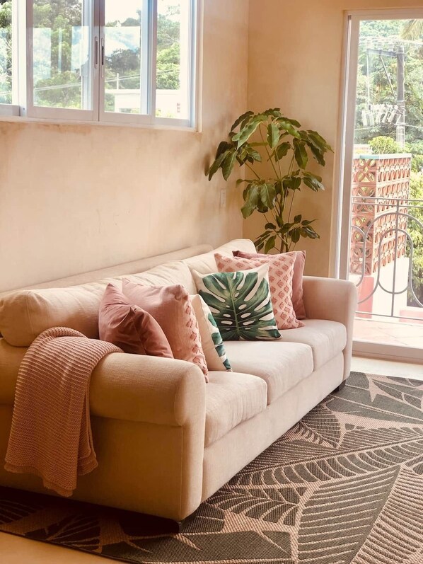 Een beige bank met een gebreide deken en meerdere kussens, een grote groene plant en een tapijt met een patroon in een kamer met grote ramen en een balkon.