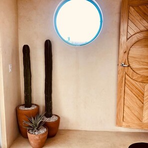 Ein Zimmer mit einem runden Dachfenster, zwei hohe Kakteen in Terrakottatöpfen und eine Holztür mit geometrischem Muster.