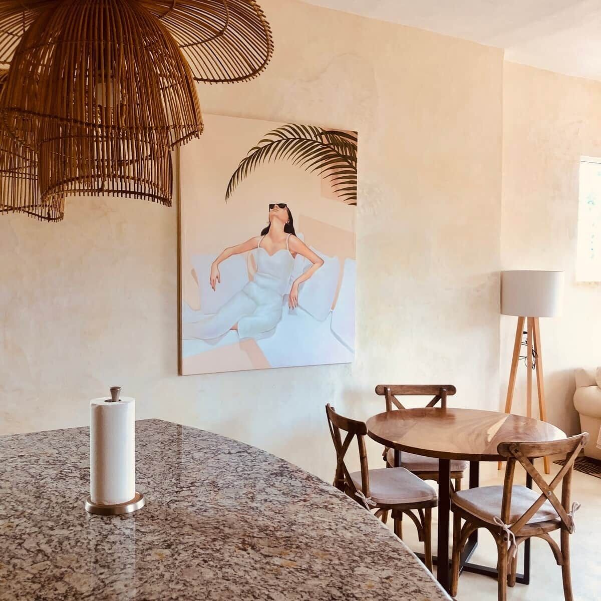 A dining area with a granite countertop, wooden chairs, a white vase, and a painting of a woman in a white dress.